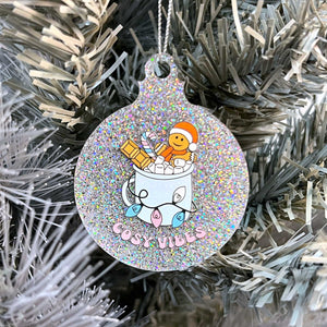 Close up of a white Christmas tree displaying a silver glitter bauble featuring retro text reading 'cosy vibes'. The image shows a mug of hot chocolate with marshmallows and a gingerbread man wearing a Santa hat. A candy cane in the mug and lights around the mug are the colours of the transgender pride flag.