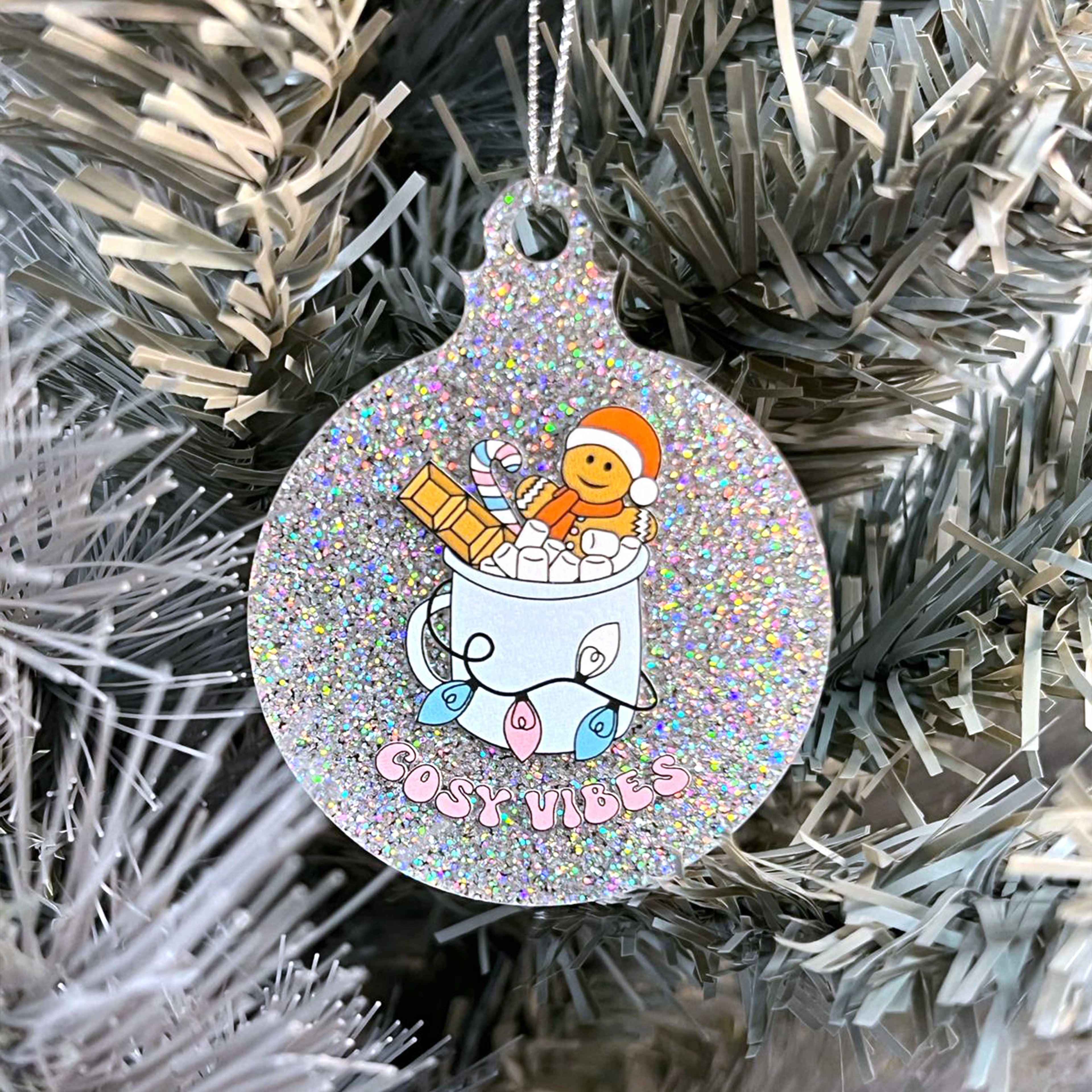 Close up of a white Christmas tree displaying a silver glitter bauble featuring retro text reading 'cosy vibes'. The image shows a mug of hot chocolate with marshmallows and a gingerbread man wearing a Santa hat. A candy cane in the mug and lights around the mug are the colours of the transgender pride flag.