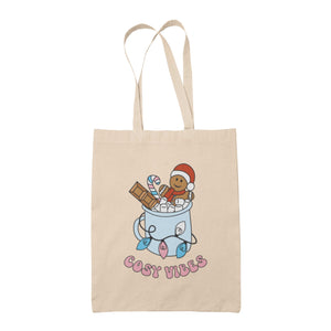 Natural tote bag featuring retro text reading 'cosy vibes'. The image shows a mug of hot chocolate with marshmallows and a gingerbread man wearing a Santa hat. A candy cane in the mug and lights around the mug are the colours of the transgender pride flag.
