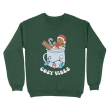 Load image into Gallery viewer, Bottle Green sweatshirt featuring retro text reading &#39;cosy vibes&#39;. The image shows a mug of hot chocolate with marshmallows and a gingerbread man wearing a Santa hat. A candy cane in the mug and lights around the mug are the colours of the transgender pride flag.