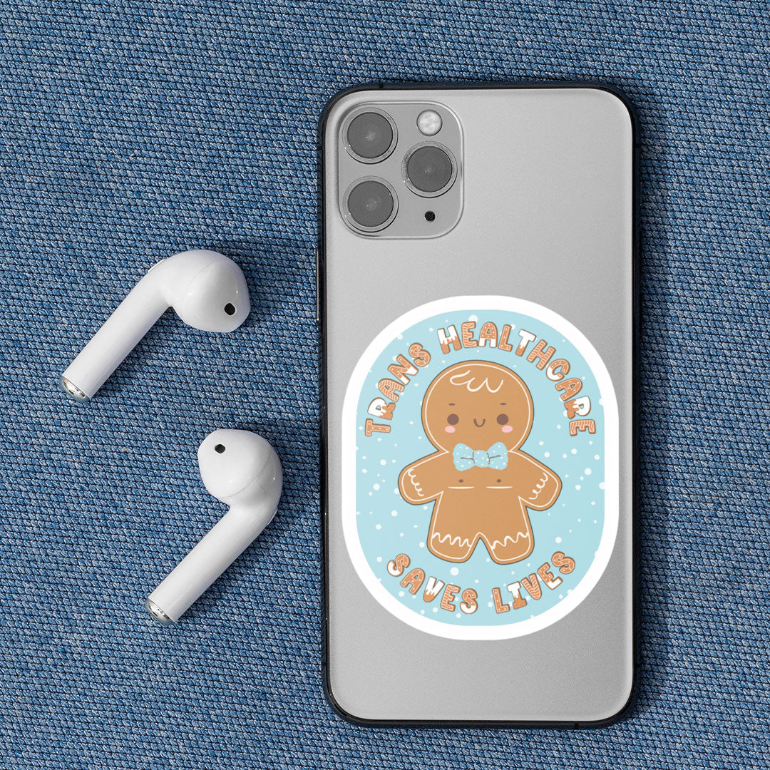 A close-up of a mobile phone with the "Trans Healthcare Saves Lives" sticker, featuring a gingerbread person wearing a bow tie and proudly displaying trans representation.