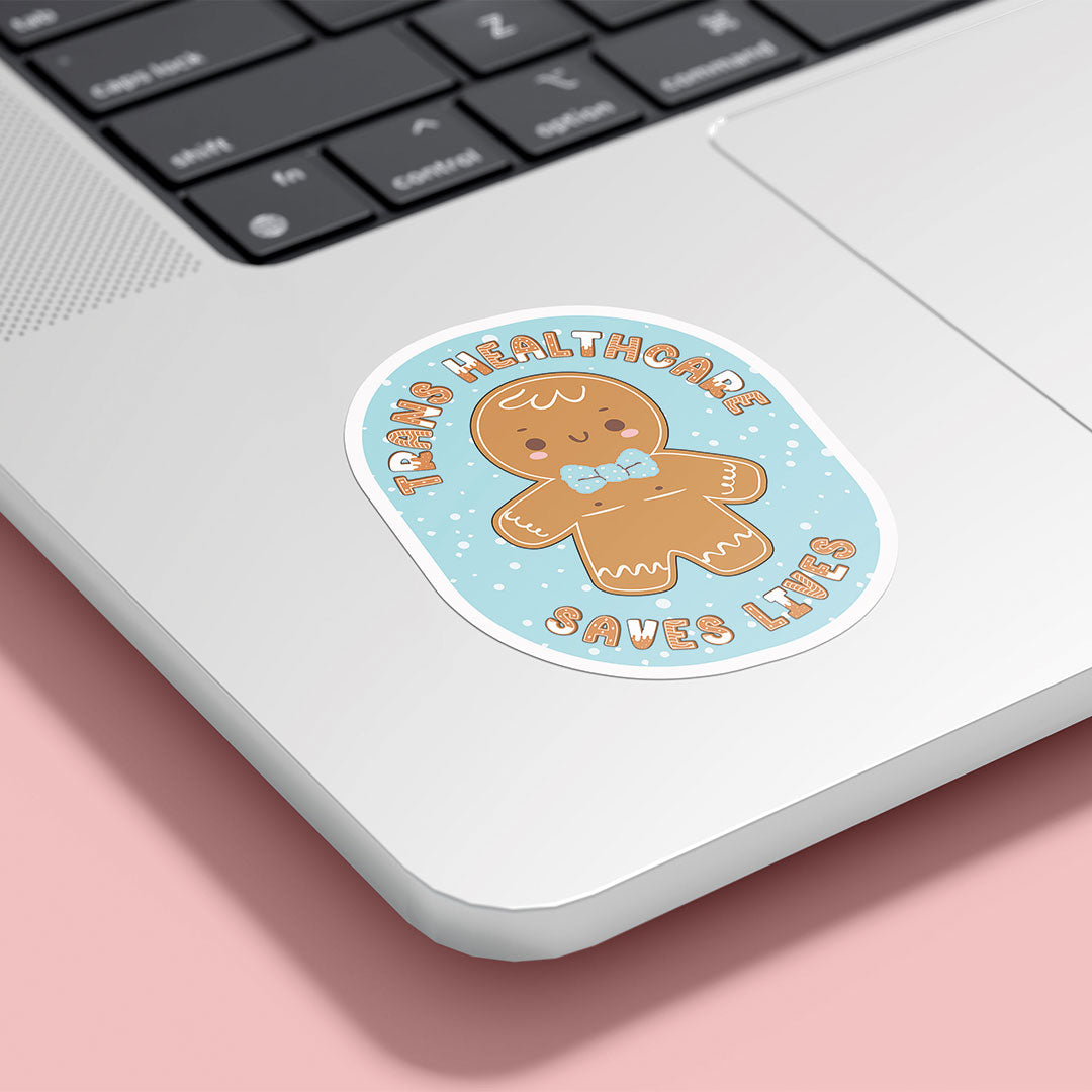 A laptop decorated with the "Trans Healthcare Saves Lives" sticker featuring a gingerbread character with top surgery scars and a cute bow tie, promoting trans healthcare awareness.