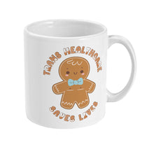 Load image into Gallery viewer, Side view of a white mug featuring the full &quot;Trans Healthcare Saves Lives&quot; design, showing a gingerbread figure with a blue bow tie and the full phrase surrounding it in a circular shape.