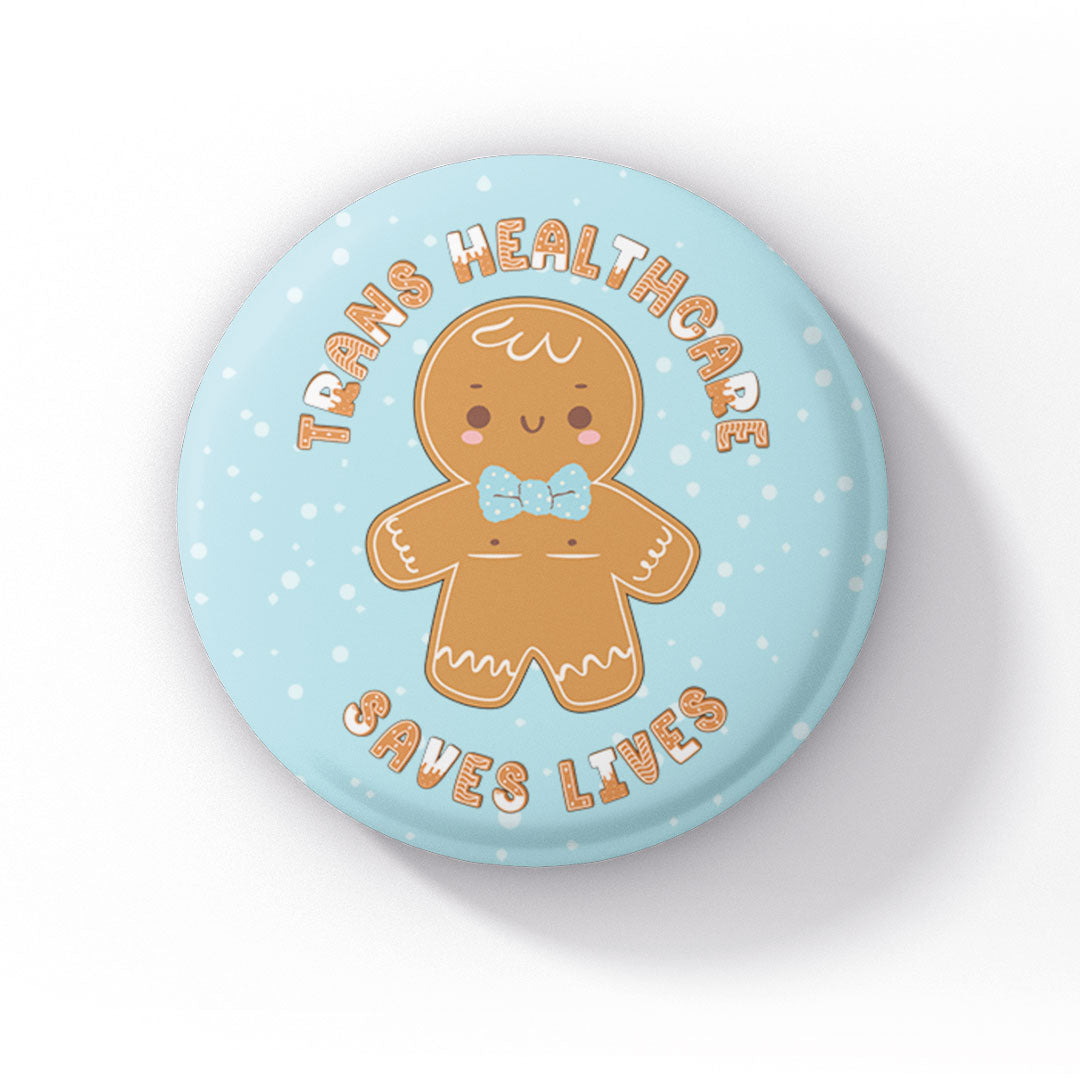 A close-up of a pastel blue 38mm badge, featuring a smiling gingerbread person with top surgery scars and a light blue bowtie. The text "Trans Healthcare Saves Lives" wraps around the design in a playful font.