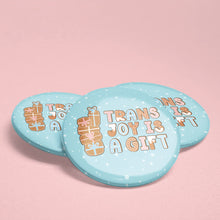 Load image into Gallery viewer, Image of three light blue badges with gingerbread-themed design that reads &quot;Trans Joy is a Gift&quot; on a soft pink background, with small present icons as part of the design.