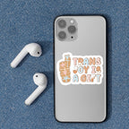 The "Trans Joy Is A Gift" sticker displayed on the back of a grey mobile phone alongside wireless earbuds on a textured blue background.