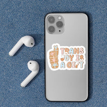 Load image into Gallery viewer, The &quot;Trans Joy Is A Gift&quot; sticker displayed on the back of a grey mobile phone alongside wireless earbuds on a textured blue background.