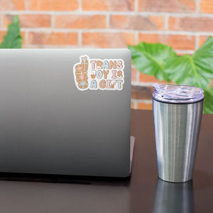 A "Trans Joy Is A Gift" sticker placed on the upper corner of a grey laptop, next to a metallic travel mug, with a brick wall and green plant in the background.