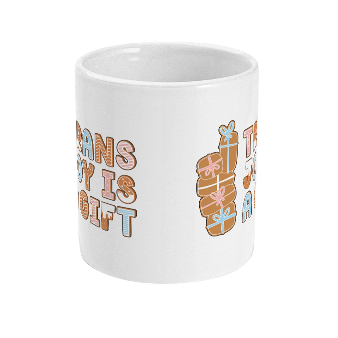 Front view of a white mug with the design "Trans Joy is a Gift" written in festive gingerbread-style lettering, accompanied by stacked gingerbread presents.