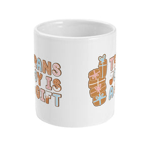 Front view of a white mug with the design "Trans Joy is a Gift" written in festive gingerbread-style lettering, accompanied by stacked gingerbread presents.