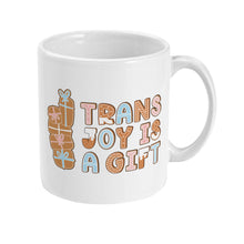 Load image into Gallery viewer, Right side view of a white mug displaying the &quot;Trans Joy is a Gift&quot; design in gingerbread-style letters with a stack of decorated gifts on the side.