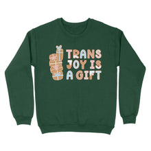 Load image into Gallery viewer, Bottle green sweatshirt featuring the text &#39;Trans Joy Is A Gift&#39; in gingerbread style, with stacked gift illustrations on the left. The design includes pastel pink, blue, and white accents.