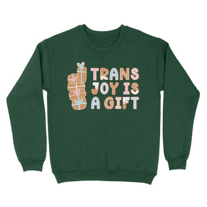 Bottle green sweatshirt featuring the text 'Trans Joy Is A Gift' in gingerbread style, with stacked gift illustrations on the left. The design includes pastel pink, blue, and white accents.