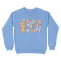 Cornflower blue sweatshirt featuring the phrase 'Trans Joy Is A Gift' styled like gingerbread cookies, with a stack of gifts to the left in pastel pink, blue, and white hues.