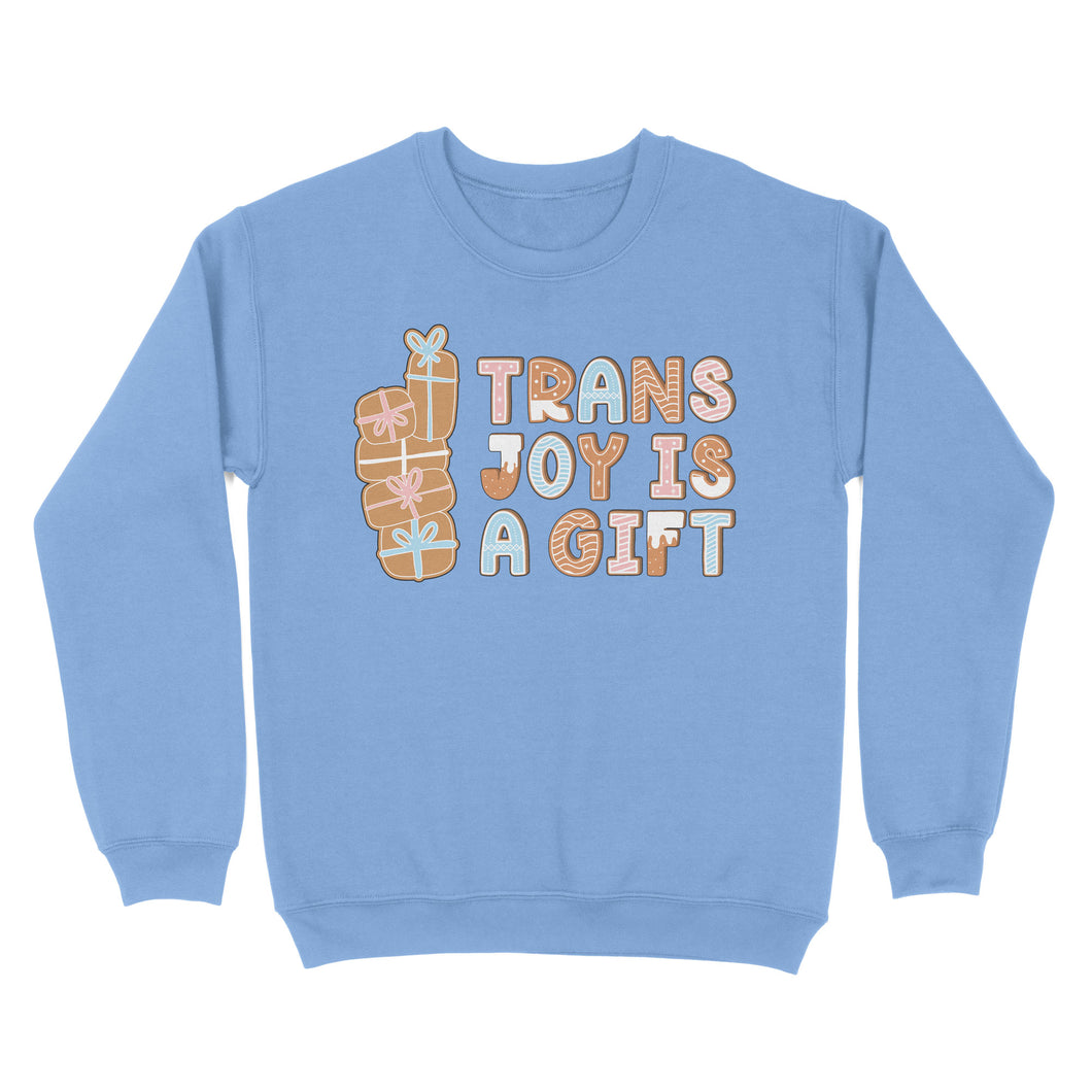 Cornflower blue sweatshirt featuring the phrase 'Trans Joy Is A Gift' styled like gingerbread cookies, with a stack of gifts to the left in pastel pink, blue, and white hues.