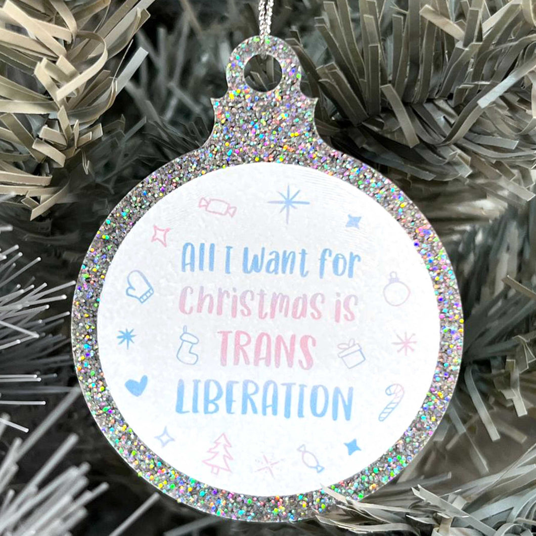 Close up of a white Christmas tree displaying a silver glitter bauble featuring the text All I Want for Christmas is TRANS LIBERATION surrounded by Christmas icons such as candy canes and stockings, all in the colours of the transgender flag; pink, blue, and white.