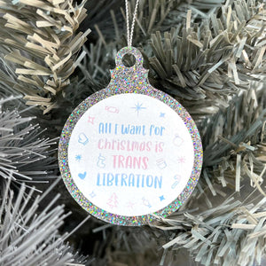 Close up of a white Christmas tree displaying a silver glitter bauble featuring the text All I Want for Christmas is TRANS LIBERATION surrounded by Christmas icons such as candy canes and stockings, all in the colours of the transgender flag; pink, blue, and white.