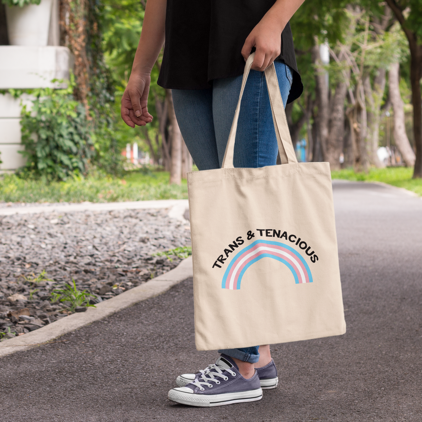 Natural cotton tote bag with “Trans & Tenacious” text and a trans pride rainbow, held at the side by a person on a park path.