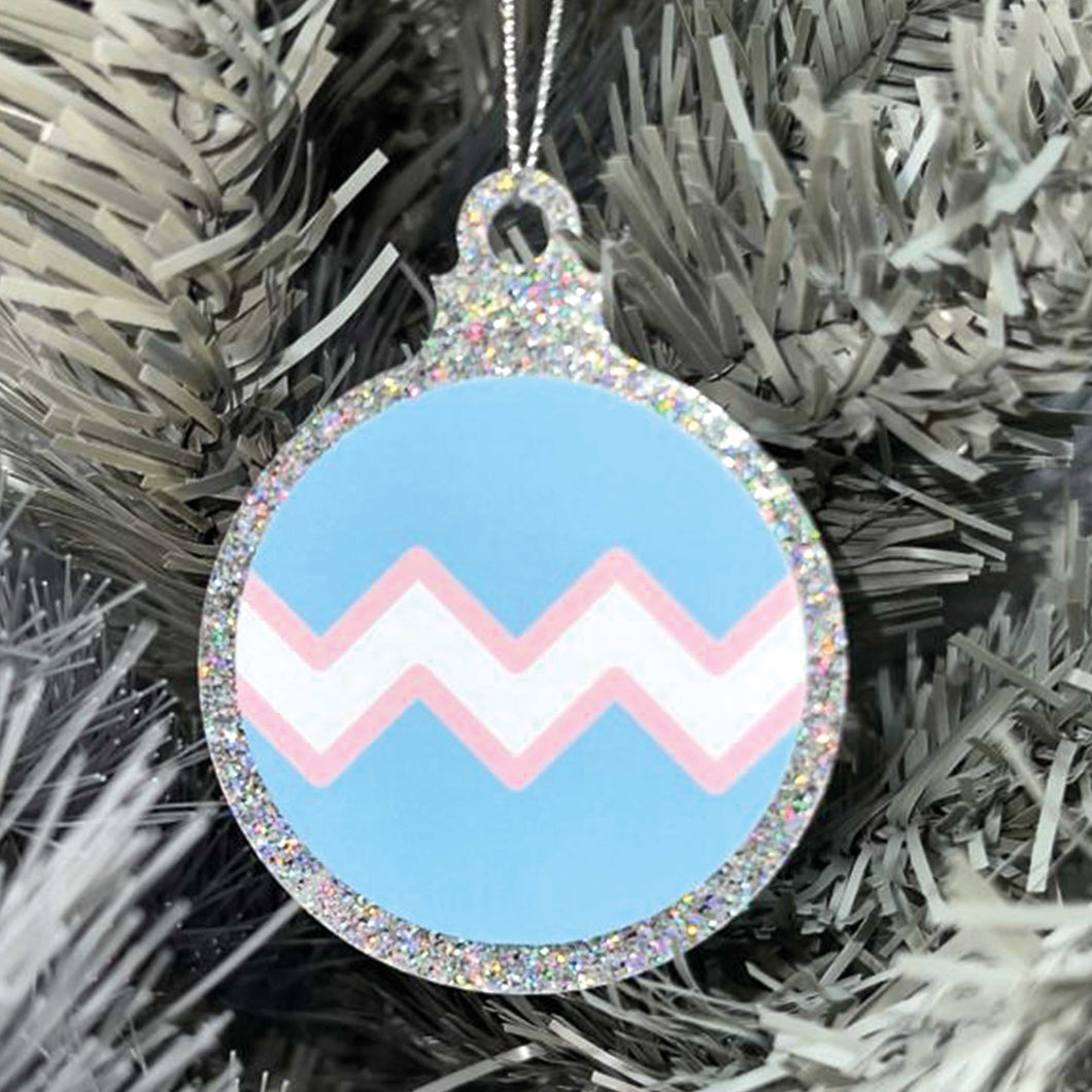 Close up of a white Christmas tree displaying a silver glitter bauble featuring a classic zig zag design in the colours of the transgender pride flag.