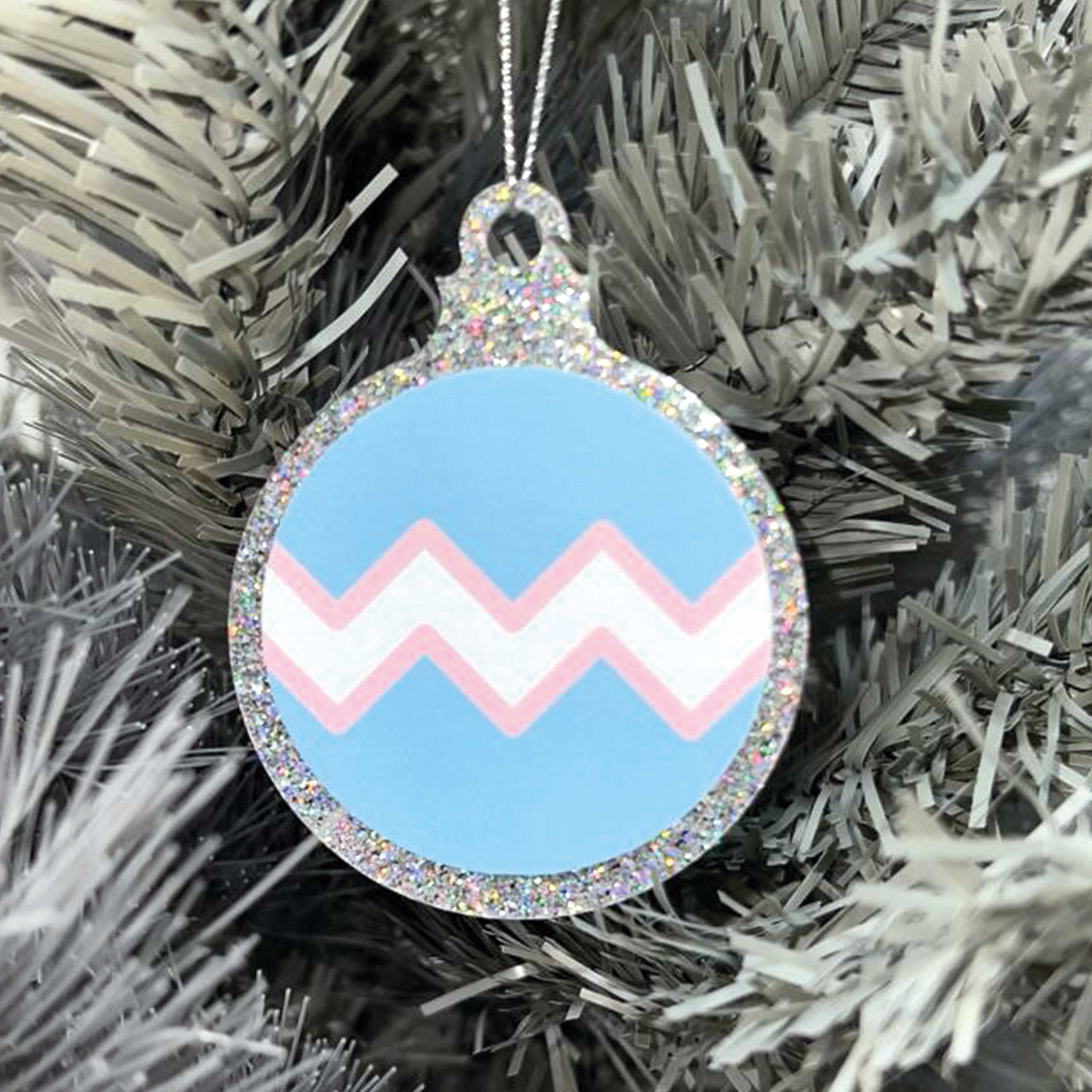 Close up of a white Christmas tree displaying a silver glitter bauble featuring a classic zig zag design in the colours of the transgender pride flag.