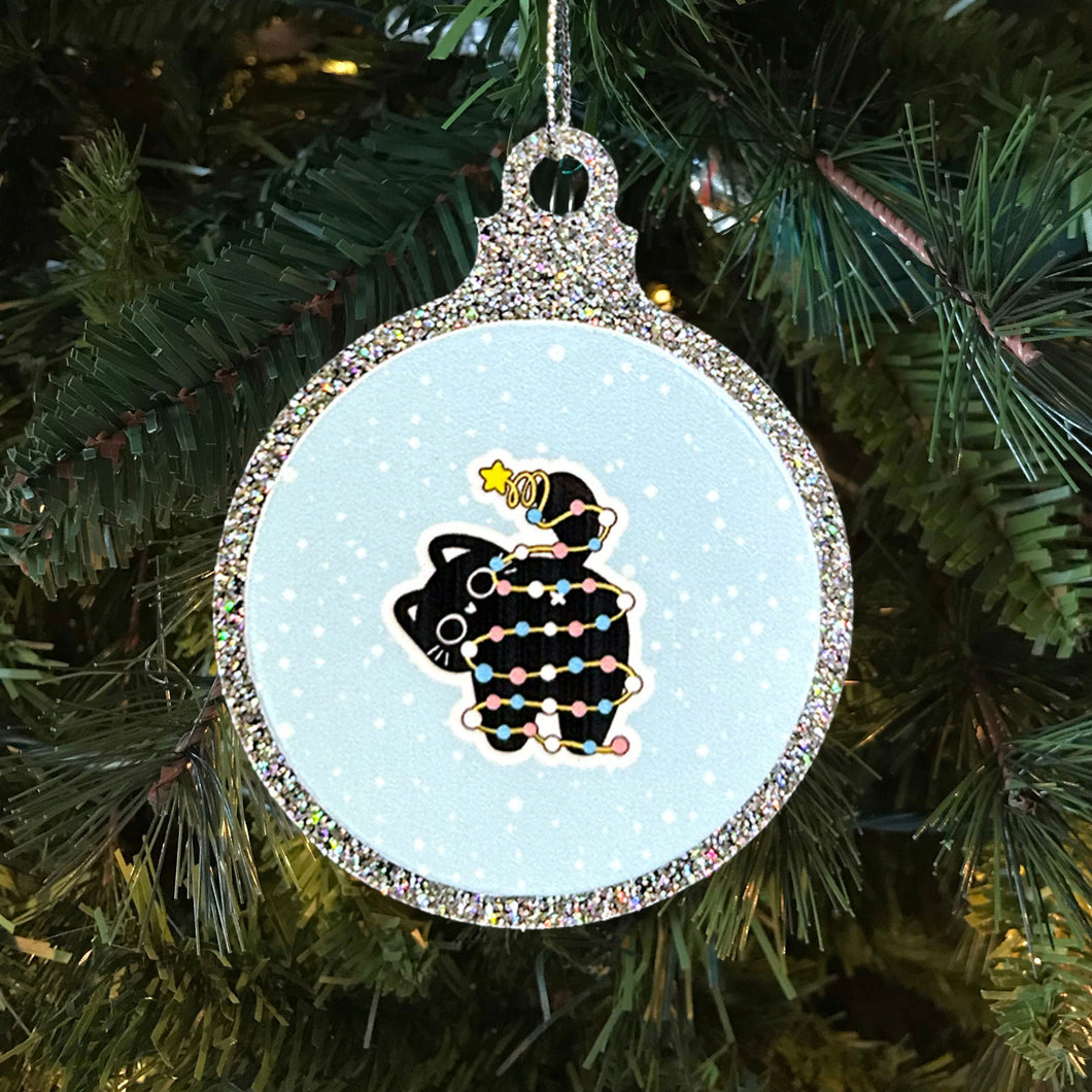 A glittery Christmas bauble featuring a black cat wrapped in fairy lights in the colours of the transgender pride flag. The cat sits cheerfully with a star on its tail, set against a snowy backdrop on a festive tree.