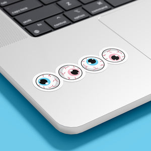 Four circular stickers with eyeball designs are placed on the bottom corner of a silver laptop. The irises are blue, pink, white, and pink, representing the transgender pride flag.