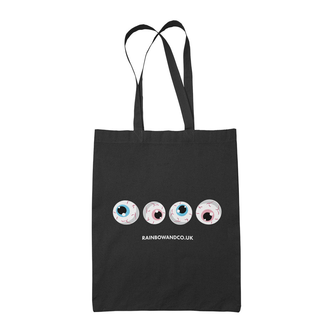 A black tote bag featuring four cartoonish eyeballs arranged in a row. Each eyeball has a different coloured iris, representing the transgender flag: one with a light blue iris, one with a pink iris, one with another light blue iris, and one with another light pink iris. Below the eyeballs, the text reads "RAINBOWANDCO.UK".