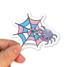 Load image into Gallery viewer, A hand holding a cute spider sticker with a transgender pride web design. The web is coloured with light blue, pink, and white segments, symbolising the transgender flag.