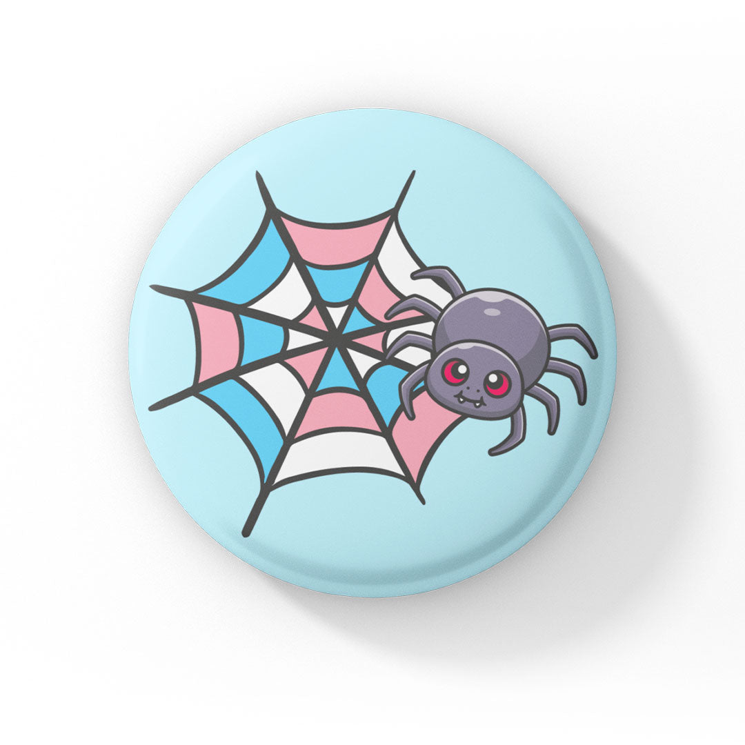 A circular badge showcasing a cute grey spider with pink eyes on a web coloured in blue, pink, and white, representing the transgender pride flag. The badge has a light blue background.
