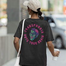 Load image into Gallery viewer, A person wearing a dark heather grey t-shirt with a bold Halloween-themed design on the back. The design features a green zombie head with an exposed pink brain and a terrified expression, surrounded by the phrase &quot;Transphobia Rots Your Brain&quot; in dripping pink letters. The person is walking outdoors, holding a white takeaway cup in one hand and wearing a light-coloured baseball cap and hoop earrings. The background shows a blurred urban setting with a parked car and buildings.