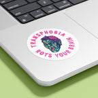 A circular sticker with the text "TRANSPHOBIA ROTS YOUR BRAIN" in bold, pink, jagged letters is placed on the bottom corner of a silver laptop. The sticker features a graphic of a zombie-like face with green skin, red eyes, and an exposed pink brain, designed to convey an anti-transphobia message. The laptop is set against a light green background.