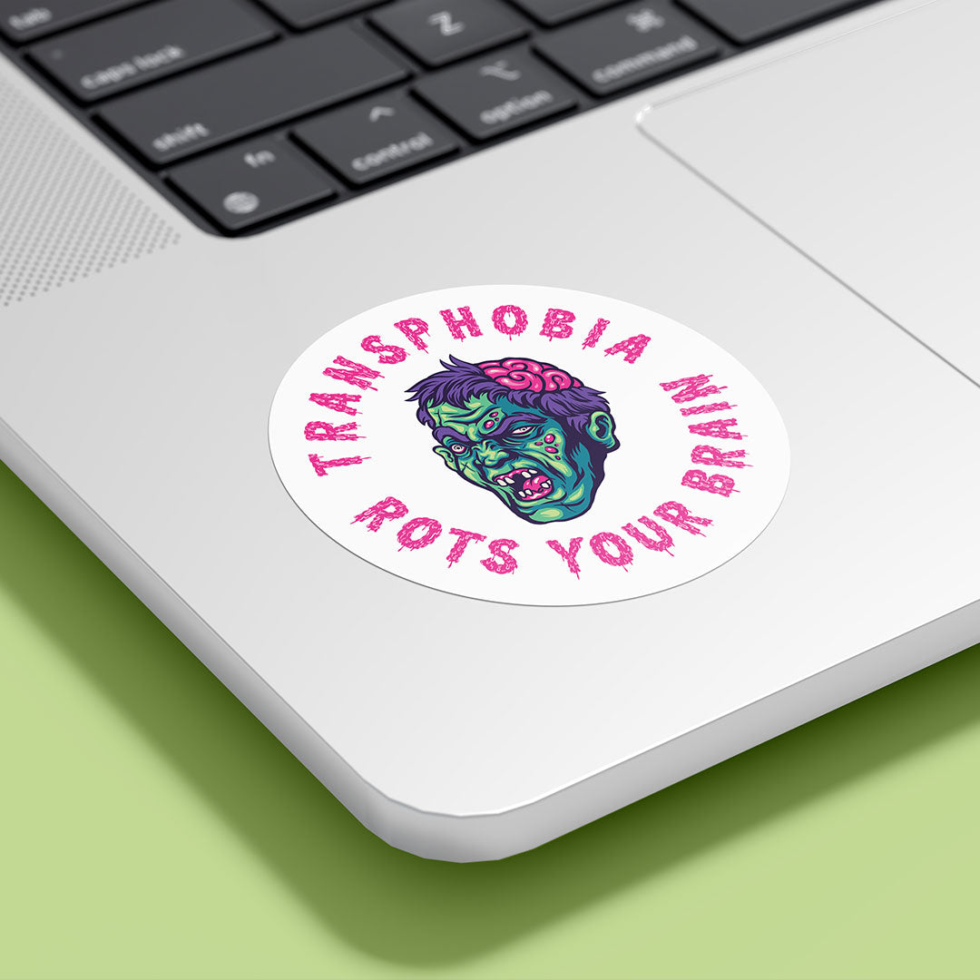 A circular sticker with the text "TRANSPHOBIA ROTS YOUR BRAIN" in bold, pink, jagged letters is placed on the bottom corner of a silver laptop. The sticker features a graphic of a zombie-like face with green skin, red eyes, and an exposed pink brain, designed to convey an anti-transphobia message. The laptop is set against a light green background.