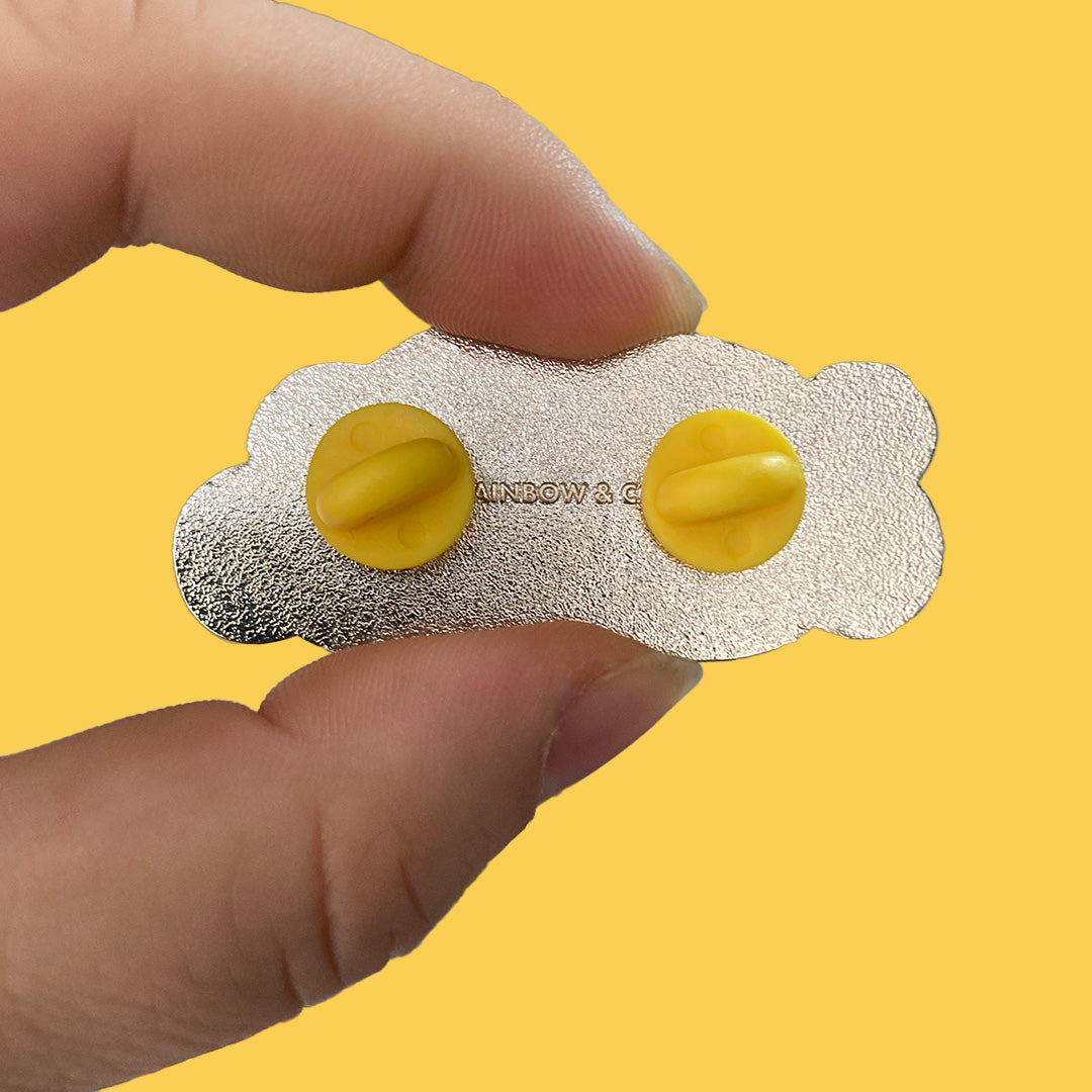 Close up of hand showing the back of a We Are Everywhere enamel pin. It has two yellow rubber clutches and is stamped with Rainbow & Co