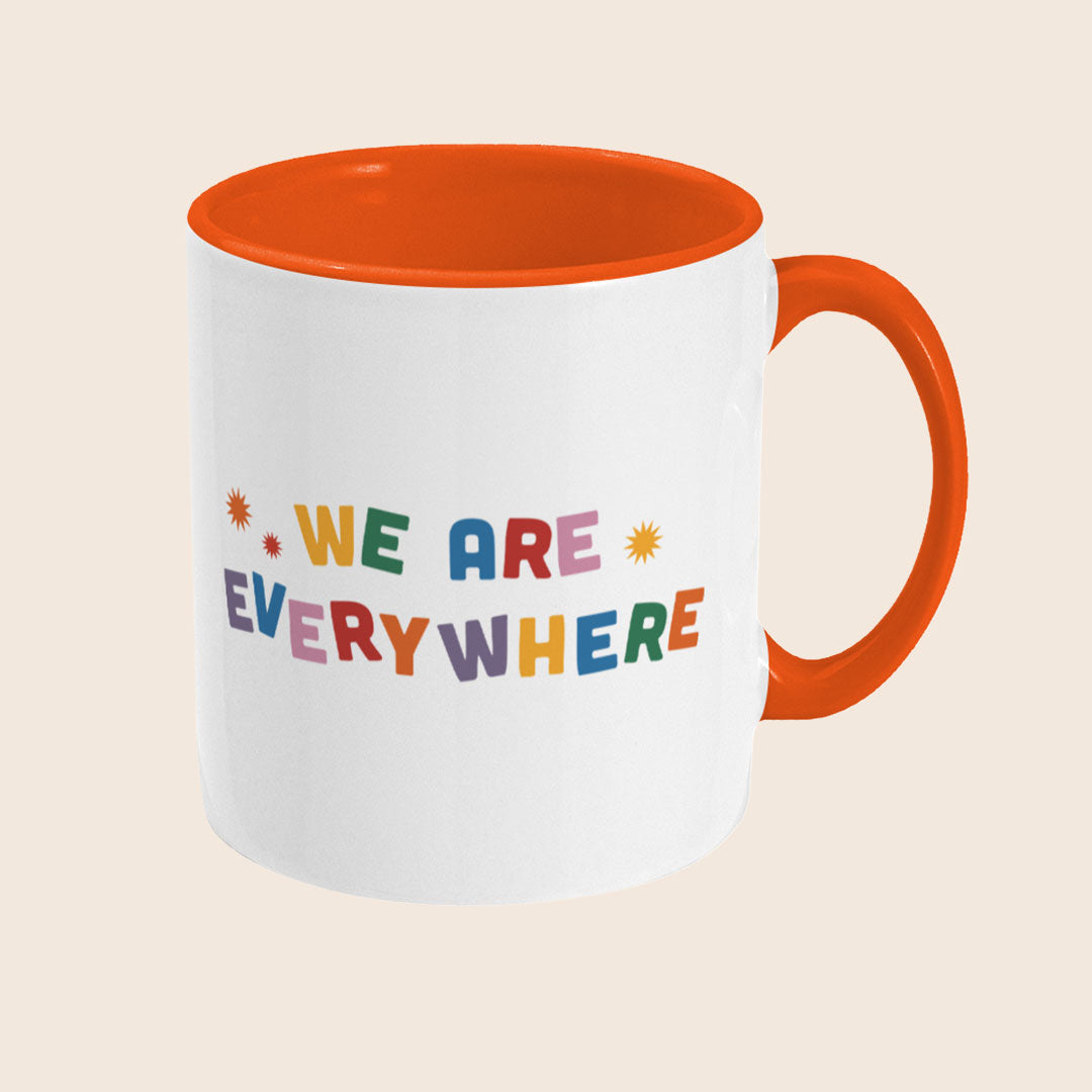 Angled view of a white ceramic mug with an orange handle and interior, displaying the colourful “We Are Everywhere” text from the opposite side.