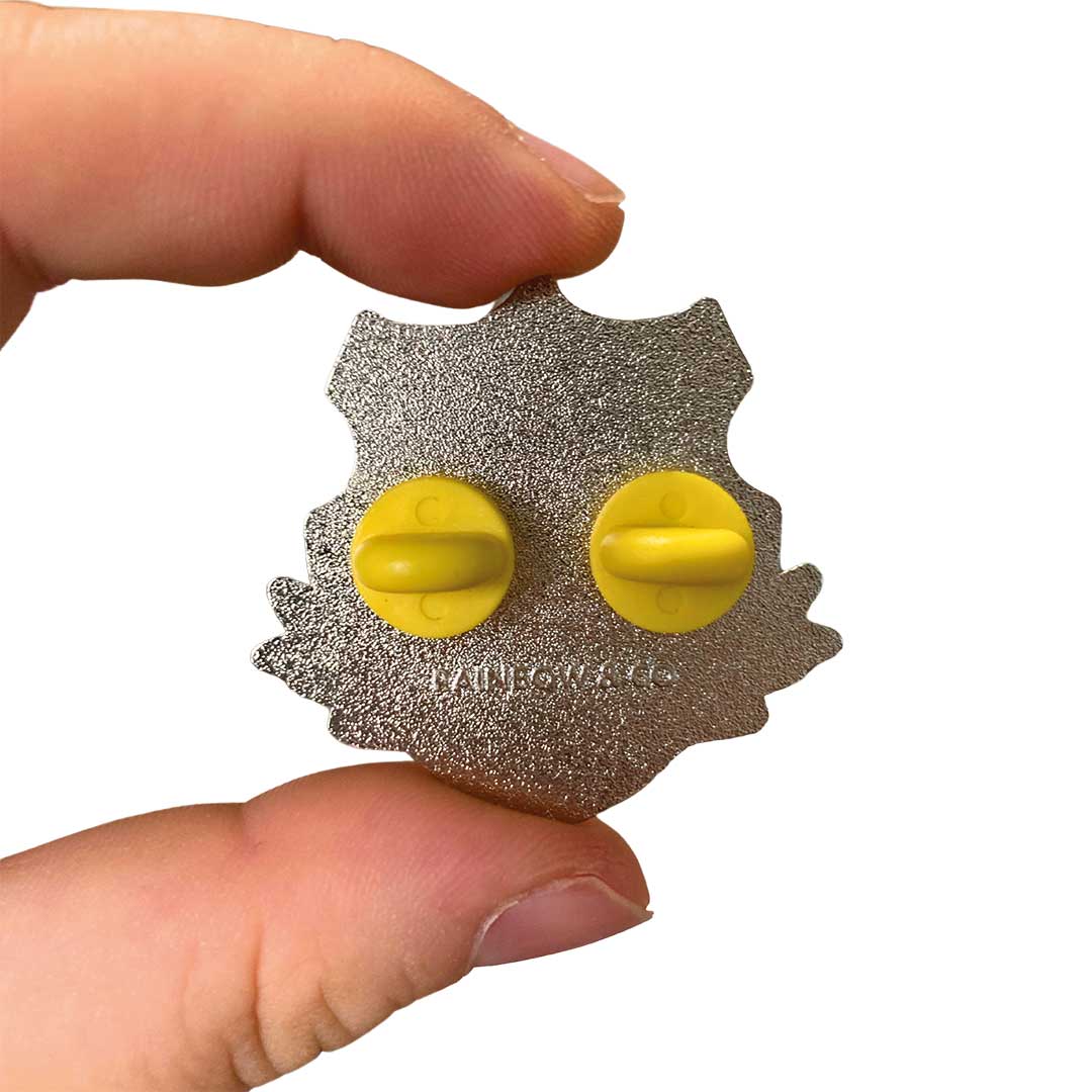 White hand holding up the reverse of a silver enamel pin in the shape of a shield. The pin has two yellow rubber backs and is stamped with RAINBOW & CO