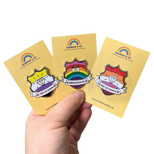 White hand holding up three yellow backing cards each showing a different shield shaped enamel pin; They of Honor, Pridesmate, and Gay of Honor. The pins are a part of the Rainbow & Co Wedding Collection.