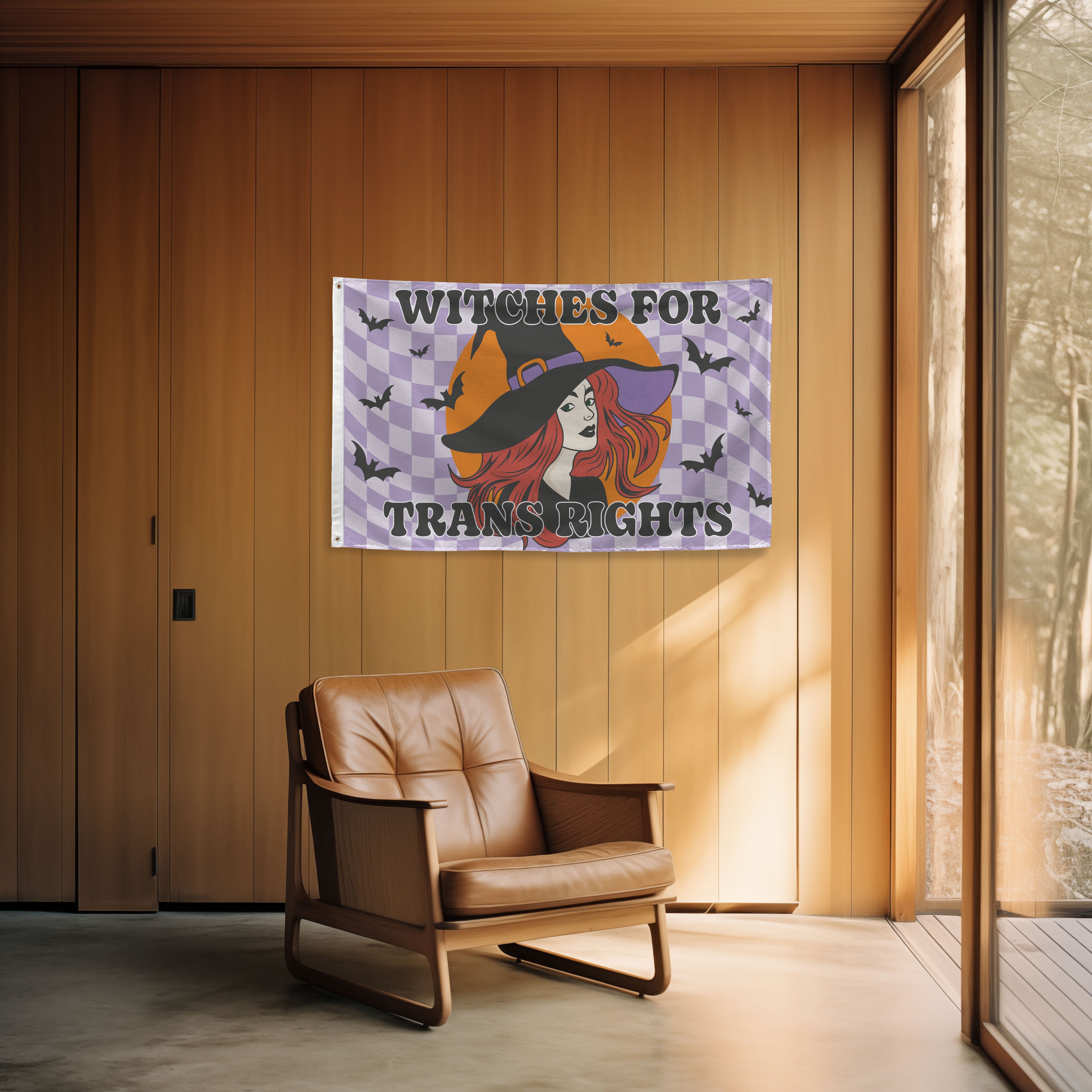 Flag with witch design and text 'Witches for Trans Rights' hanging on a wooden wall above a brown leather chair.