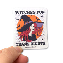 Load image into Gallery viewer, Gloss sticker being held between a thumb and forefinger. The sticker features the slogan &#39;Witches For Trans Rights&#39; alongside an image of a witch surrounded by bats.