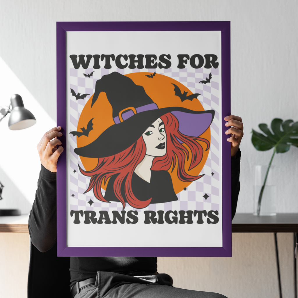 Seated person holding a purple-framed “Witches for Trans Rights” art print in front of their face. The design features a red-haired witch in a black hat, an orange moon, bats and a lilac chequerboard background.