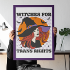 Seated person holding a purple-framed “Witches for Trans Rights” art print in front of their face. The design features a red-haired witch in a black hat, an orange moon, bats and a lilac chequerboard background.