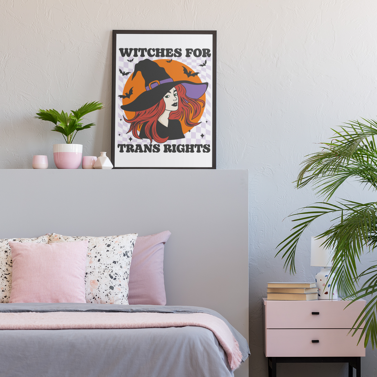 Black-framed “Witches for Trans Rights” art print featuring a red-haired witch, orange moon and bats, displayed above a bed with grey bedding and pink cushions, beside plants and ornaments.