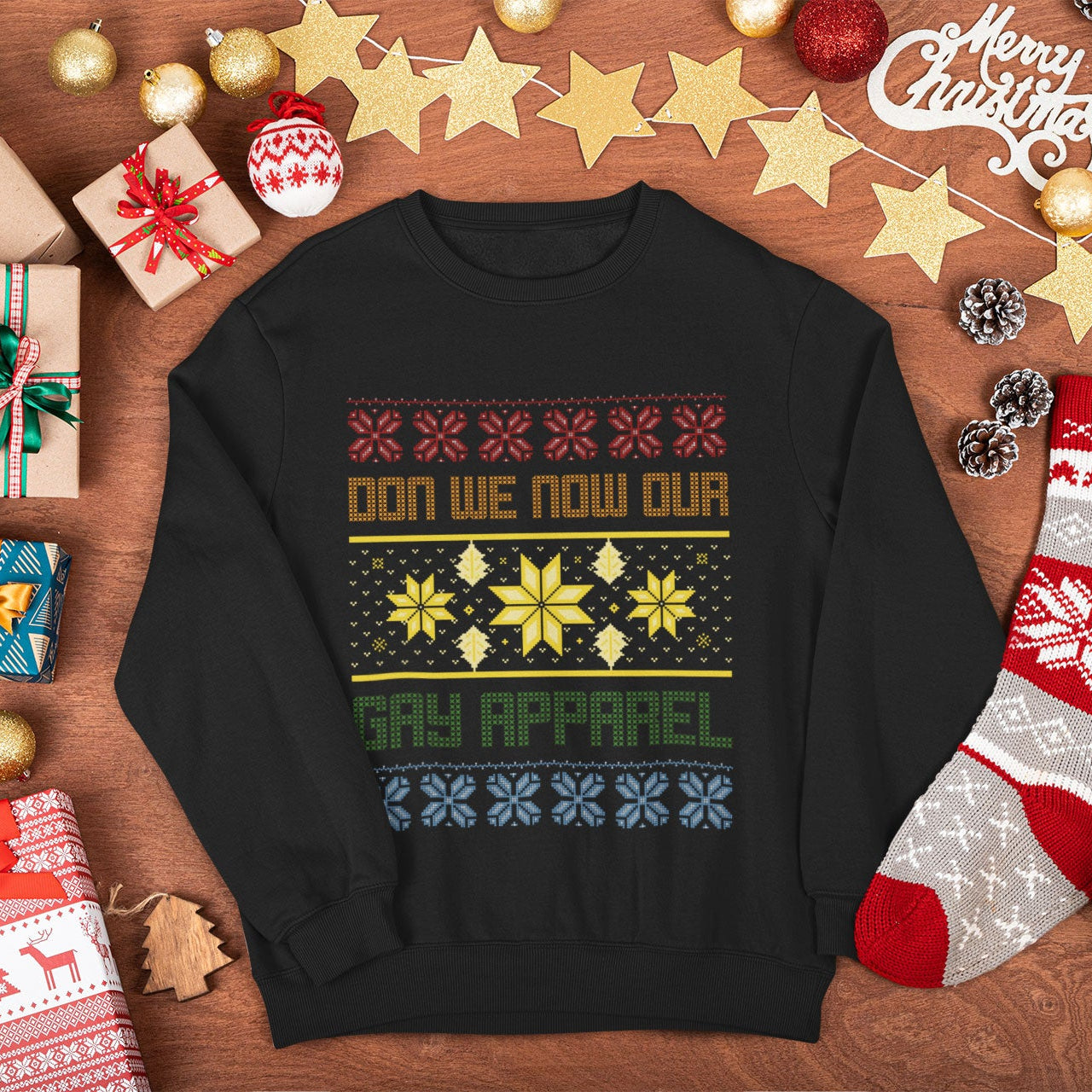 A black crewneck sweatshirt with a colorful Christmas-themed graphic reading 'DON WE NOW OUR GAY APPAREL' surrounded by holiday decorations like ornaments, gifts, pinecones, and a stocking on a wooden surface.