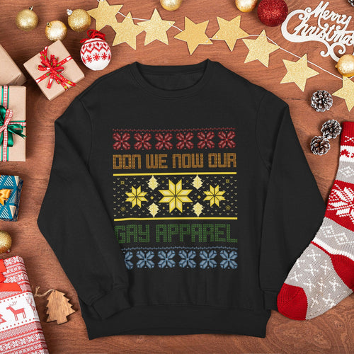 A black crewneck sweatshirt with a colorful Christmas-themed graphic reading 'DON WE NOW OUR GAY APPAREL' surrounded by holiday decorations like ornaments, gifts, pinecones, and a stocking on a wooden surface.