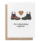 White greeting card featuring two Chelsea-style boots facing each other with a heart above and the words "Our soles belong together." with a brown kraft envelope behind.
