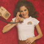 Retro style photo of a young woman lying on her back on a red plush carpet, holding a retro style telephone in her hand. She is wearing a Gay is Proud pride t shirt from Rainbow & Co.