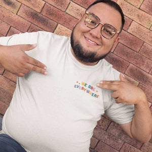 A smiling man wearing large glasses pointing at the shirt he is wearing which displays the phrase 'we are everywhere'.